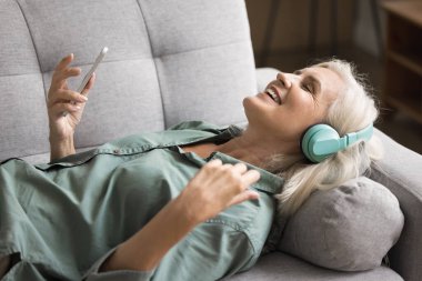 Gülümseyen 65 'li kadınlar kablosuz kulaklık takıyor. Kanepeye uzanmış, akıllı telefon tutuyor ve çevrimiçi dijital yayın hizmetleri uygulaması kullanarak en sevdiği şarkıyı dinliyor. Hobi, müzik aşığı, modern teknoloji