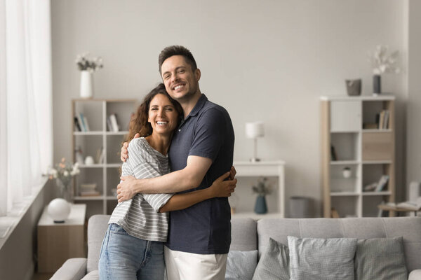Cheerful attractive young married couple of new homeowners portrait. Happy husband and wife standing in modern home interior, hugging, smiling, laughing, looking at camera