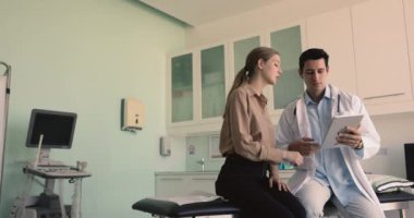 Beyaz önlüklü, elinde dijital tabletle klinik muayene odasında tıbbi tedaviyi açıklayan erkek pratisyen doktorla konuşan güzel bir kadın hasta. Tıp, modern teknoloji.