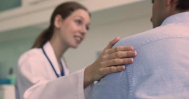 Genç kadın pratisyen doktor, erkeğin omzuna dokunur, onu rahatlatır, empati kurar, destekleyici konuşma yapar, cesaret verir, psikolojik yardım sağlar ve iyileşmesi için umut verir.