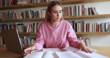 Odaklanmış zeki beyaz kız kütüphanede kitap yığınları okuyor, kitap rafları arkasında, notlar yazıyor, rapor için özet yapıyor, araştırma çalışması yapıyor, laptopun başında oturuyor.
