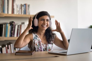 Çalışmak için müzikle heyecanlanan mutlu Hintli genç kız büyük kulaklıklar takıyor, dizüstü bilgisayar başında oturuyor, başka tarafa bakıyor, gülümsüyor, müzik sesinden hoşlanıyor, çevrimiçi iletişim kuruyor.