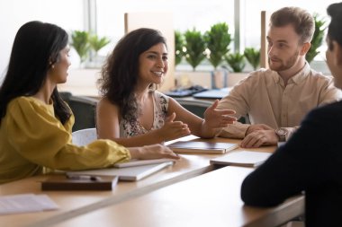 Mutlu Hintli proje yöneticisi kadın beyin fırtınası toplantısında iş arkadaşlarıyla konuşuyor, takım çalışması için yaratıcı bir plan sunuyor, gülümsüyor, konuşuyor, el ele tutuşuyor, büyük bir masada oturuyor.