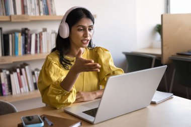 Ciddi genç Hintli internet öğretmeni internet, online ders veriyor, kulaklık takıyor, bilgisayar başında konuşuyor, kütüphaneden yayın yapıyor, ekranda konuşuyor.