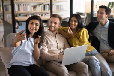 Çeşitli ofis arkadaşları birlikte akıllı telefondan selfie çekiyorlar, parmak kalp işareti gösteriyorlar, modern iş ortaklığında boş vakitlerin tadını çıkarıyorlar, fotoğraf çekiyorlar, gülümsüyorlar.