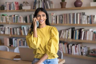 Halk kütüphanesinde cep telefonuyla konuşan, arka planda dizüstü bilgisayarı ve kitaplığı olan, yüzünü çeviren, gülümseyen, gülen, konuşan mutlu genç Hintli üniversiteli kız.