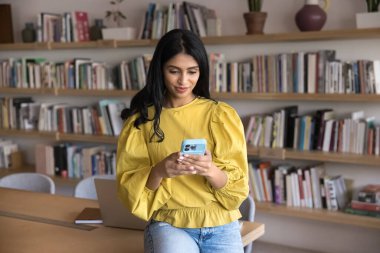 Odaklanmış genç Hintli yönetici kadın akıllı telefona yazıyor, ofis kütüphanesindeki çalışma masasına yaslanıyor, modern çalışma alanlarında iş internet iletişimi için cep telefonu kullanıyor.