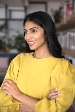 Kollarını kütüphanede kavuşturmuş, dişlek gülümsemesiyle yüzünü çevirmiş, dikey portre için poz veren, eğitimden zevk alan, okumaktan zevk alan güzel Hintli genç bir kız öğrenci.