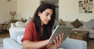 Çevrimiçi alışveriş için dijital tablet kullanan gülümseyen Latin bir kadın, arkadaşıyla uzaktan iletişim kuran, sipariş veren, müşteriyle iletişim kuran, evden telgraf çeken. Modern teknoloji, internet ve e-randevu uygulaması kullanımı