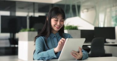 Genç gülümseyen Çinli iş kadını, modern tablet cihazını kullanarak çalışan ofis çalışanı, çalışma odasında rahat bir şekilde oturup şirket müşterilerine mektuplar gönderen, posta kutusunu kontrol eden,