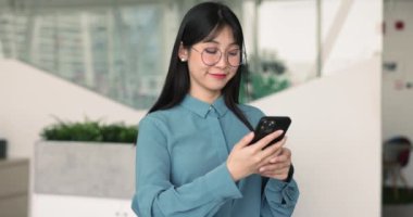 Gülümseyen Asyalı kadın ofiste akıllı telefon kullanarak gözlük takıyor, e-posta yoluyla müşteriyle iletişim kuruyor, yeni konforlu zaman yönetimi uygulamasının keyfini çıkarıyor, iş arkadaşlarıyla sohbet ediyor, yapılacaklar listesi yaratıyor, mesajları kontrol ediyor