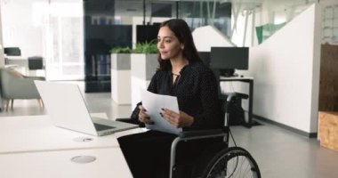 Tekerlekli sandalyede oturan genç bir kadın erişilebilirlik için, dizüstü bilgisayar kullanarak, belgeleri inceleyerek, ofiste verimli bir şekilde çalışıyor. Görevleri arasında dijital ve kağıt tabanlı sorumluluklar bulunuyor.