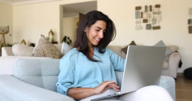 Dizüstü bilgisayarda çalışan çekici bir kadın rahat bir oturma odasında rahat bir şekilde oturuyor, uzaktan çalışıyor, profesyonel işleri yürütüyor. E-servis satın alma ve tele-çalışma için modern teknoloji kullanımı