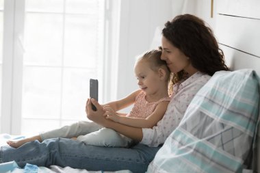 Mutlu güzel kız annesinin kucağında oturuyor, internet uygulamasını cep telefonunda kullanıyor, kablosuz iletişim teknolojisi kullanıyor, selfie çekiyor, aile içi konuşmalar yapıyor, evde dinleniyor.