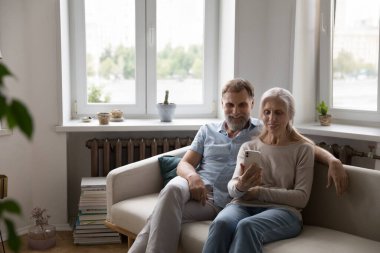 Mature couple sit on sofa staring at smart phone screen having video call remote conversation with grown up children or relative living abroad. Older gen use modern tech, virtual meeting event concept clipart