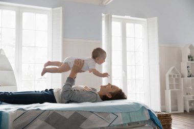 Neşeli genç anne kucağında çocuk, tatlı pozitif bebeği havaya kaldırıyor, yatağa uzanıyor, gülümsüyor, gülüyor, bebekle uçak oynuyor, boş zamanların tadını çıkarıyor, annelik yapıyor.