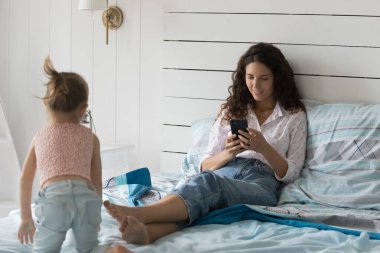Pozitif anne oturuyor, yatakta dinleniyor, internet bağlantısı için cep telefonu kullanıyor, ekommerce uygulaması ile online alışveriş yapıyor. Annesinin oyuncağıyla oynayan aktif bir kız çocuğu.