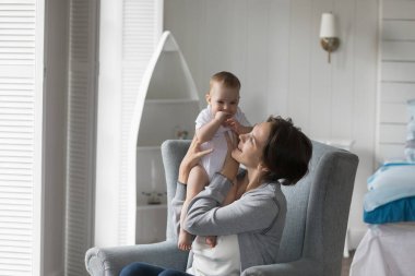 Neşeli, genç bir anne, tapılası bir bebeğe sarılıyor, kucağında küçük bir çocuk, koltuğunda oturuyor, gülümsüyor, gülüyor, mutlu anneliğin tadını çıkarıyor, annelik izninde.