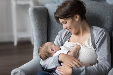 Mutlu genç anne, bebeği emziriyor. Sevgi, şefkat, bağlılık, şefkat, gülümseme, kucağında çocuk olan sevimli, uykulu çocuğa bakıyor. Yeni anne anneliğin tadını çıkarıyor.