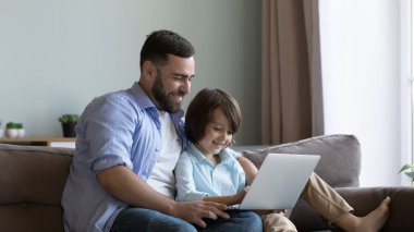 Neşeli bin yıllık baba ve mutlu nesil Z oğlu dizüstü bilgisayarı evde birlikte kullanıyor, daktilo ediyor, öğrenme uygulaması okuyor, internette oyun oynuyor, alışveriş yapıyor, dijital aygıtı internet iletişimi için kullanıyor.