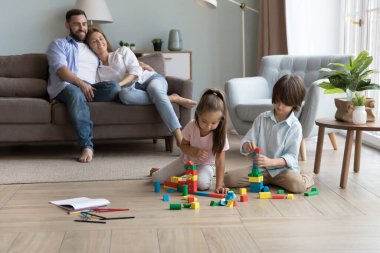 Oyuncak bloklarla oynayan iki odaklanmış sakin çocuk, sıcak ısıtma tabanında küpler, kuleler inşa eden, renkli kaleler inşa eden. Bir çift ebeveyn kanepede dinlenir, evde küçük çocukları izlerler.