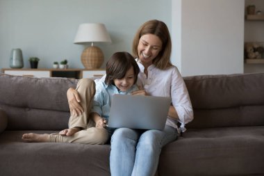 Neşeli güzel anne ve meraklı çocuk, anaokulu için online öğrenme uygulaması kullanıyor, çizgi film izliyor, video çağrısı yapıyor, kablosuz bağlantı için cihaz tutuyor, iletişim kuruyor, gülüyor.