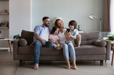 Mutlu bin yıllık ebeveynler ve iki küçük çocuk cep telefonlarını ev koltuğunda kullanıyor, sarılıyor, gülüyor, eğleniyor, eğleniyor, eğleniyor, internetten iletişim kuruyor, internet üzerinden alışveriş yapıyor.