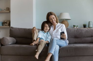Mutlu bir anne ve tatlı küçük bir oğul evde kanepede dinleniyor, akıllı telefon kullanıyor, video görüşmesinde konuşuyor, cep telefonu tutuyor, gülümsüyor, gülüyor. Çevrimiçi iletişim, aile kavramı