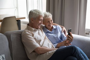 Olgun bir çift akıllı telefonuyla kanepeye oturur, cihaz ekranına bakar, cep telefonuyla selfie çektirir, online videolar izler, video görüşmelerini mutlu eder, evdeki modern kablosuz teknolojiyi kullanır, eğlenceli bir konsept oluşturur.