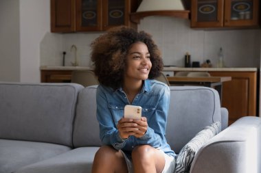 Şirin Afrikalı genç kız rahat kanepesinde dinleniyor akıllı telefonu elinde tutuyor yan bakıyor rüya gibi görünüyor, modern cihaz kullanımından dikkati dağılıyor, satın almaktan memnun olduğunu düşünüyor teknoloji kullanarak evde boş zaman geçiriyor.