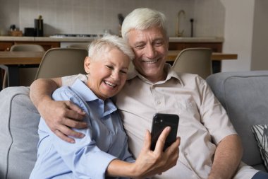 Mutlu, yaşlı ve yakışıklı bir çift kanepeye oturup akıllı telefon uygulaması yapıyor. Modern teknoloji kullanımı, yetişkin çocuklarla sanal buluşma, uzaktan iletişim, video konferans konsepti