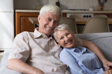 Beautiful hoary good-looking mature spouses relaxing together on comfy couch at modern home smiling staring at camera feel carefree. Good harmonic relationships, happy marriage, endless love concept