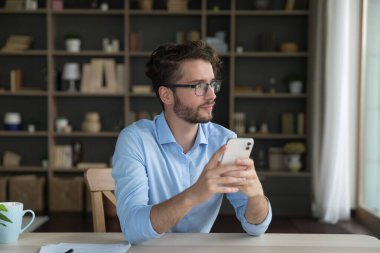 Düşünceli hayalperest iş adamı elinde akıllı telefon, çalışma masasında oturuyor, pencereye bakıyor, düşünüyor, daktilo ediyor, internette sohbet ediyor, cep telefonunda internet teknolojisini kullanıyor.