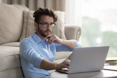 Ciddi odaklı, havalı gözlüklü, dizüstü bilgisayarda çalışan, evde oturan, iş için kablosuz teknolojiyi kullanan internetteki boş zamanlarında düşünen, çenesine dokunan serbest çalışan genç bir adam.