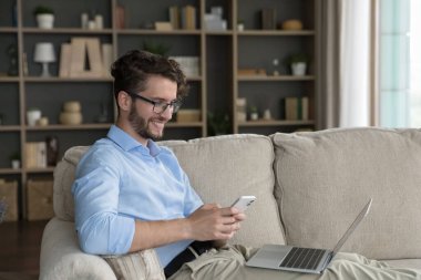 Mutlu pozitif genç girişimci evde internet teknolojisinden zevk alıyor, cep telefonu kullanıyor, iş iletişimi için dizüstü bilgisayar kullanıyor, dijital cihazlarla kanepede dinleniyor, ekrana bakıyor.