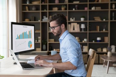 Masaüstü büyük ekranda finansal verileri analiz eden, pazarlama grafikleri, satış raporları üzerinde çalışan, iş için modern internet teknolojisini kullanan, dizüstü bilgisayarda yazan genç serbest çalışan bir iş adamı.