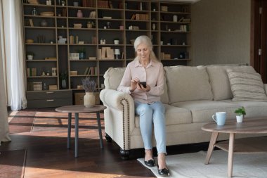 Evde akıllı telefon kullanan mutlu bir bayan, oturma odasında koltukta oturuyor, mesajlaşıyor, internette geziniyor, alışveriş için yeni bir uygulama kullanıyor, medya servisi yapıyor, gülümsüyor. Tam boy geniş çekim