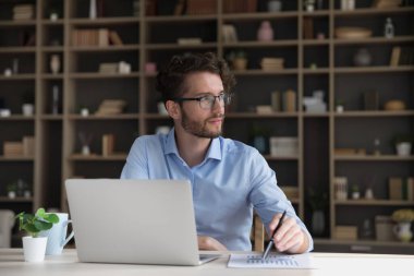 Düşünceli, genç ve ciddi bir iş adamı dizüstü bilgisayarda çalışıyor, derin düşüncelere dalıyor, kağıt pazarlama raporunu düşünüyor, interneti serbest çalışmak için kullanıyor.