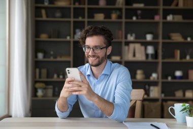Neşeli genç iş adamı uygulamayı kullanıyor, cep telefonuyla evde internet iletişimi kuruyor, masada oturuyor, daktilo yazıyor, gülümsüyor, kablosuz teknolojiden keyif alıyor.