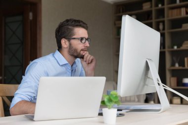 Ciddi meşgul genç serbest çalışan gözlüklü adam online proje üzerinde çalışıyor, iş istasyonu bilgisayarını kullanıyor, internet teknolojisini serbest proje için kullanıyor, büyük ekrana bakıyor, çeneye dokunuyor, düşünüyor.