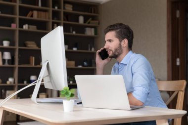 İşyeri bilgisayarında telefonla konuşan, müşteri ve müşterilerle proje tartışan, modern kablosuz internet teknolojisini iş iletişimi için kullanan meşgul genç serbest çalışan.