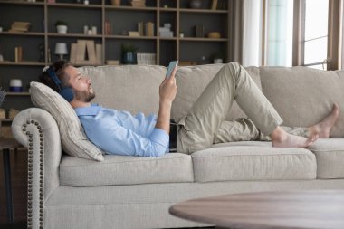 Huzurlu bin yıllık genç adam büyük kablosuz kulaklıklarıyla rahat koltukta dinleniyor, meditasyon için ortam müziği dinliyor, internetteki çalma listesini kullanıyor, sessizliğin tadını çıkarıyor, evde mola veriyor.