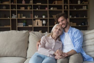 Mutlu, hayalperest, emekli anne ve yetişkin oğul yan yana oturuyor, toplantının keyfini çıkarıyor, aile zamanı, sarılıyor, el ele tutuşuyor, gülümsüyor, gülüyor, başka tarafa bakıyor.