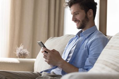 Mutlu yakışıklı genç adam interneti cep telefonuyla tarıyor, uygulamayı kullanıyor, akıllı telefondaki yazılımı kullanıyor, yumuşak koltukta oturuyor, sohbet ediyor, iyi haberler okuyor, iletişimden keyif alıyor.