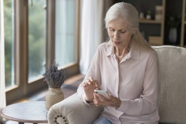 İletişim için akıllı telefonu kullanan, evde oturan, daktilo eden, sosyal medyayı karıştıran, internetten online uygulama kullanan, medya içeriğini izleyen yaşlı bir kadın.