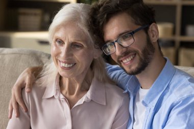 Mutlu kıdemli büyükanne ve yakışıklı torunun kucaklaşması, evde çekim yapmak için poz vermesi, koltukta oturması, başka yere bakması, gülümsemesi, gülmesi. Yaşlı anne ve yetişkin çocuk portresi