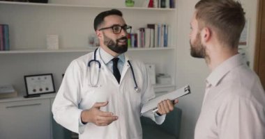 Erkek hastaya danışmanlık yapan erkek hekim, iyileşmiş hastalıklar, sağlık durumları, tokalaşma hakkında iyimser haberleri paylaşıyor. Pozitif iletişim, etkili tedavi, profesyonel tıbbi bakım sağlayıcı