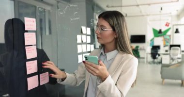 Modern akıllı telefon kontrol hatırlatıcıları kullanan iş kadını ya da cam tahta duvara yapıştırılmış notlar üzerine zamanlayıcı, yazılı bilgileri karşılaştırma, düzenli kalmak için araçlar kullanarak görevlerini yönetme
