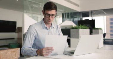 Genç odaklı işadamı ofisinde oturuyor, dizüstü bilgisayarla çalışırken belgelere bakıyor, önemli bilgileri inceliyor veya analiz ediyor, sunum hazırlıyor. Veri analizi, genel bakış