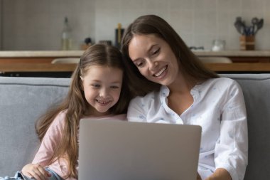 Mutlu anne ve küçük kız sanal uygulamayı dizüstü bilgisayarda kullanıyor, bilgisayarı tutuyor, birlikte oturuyor, kitap okuyor, video konferans görüşmesi yapıyor, gülümsüyor, gülüyor.